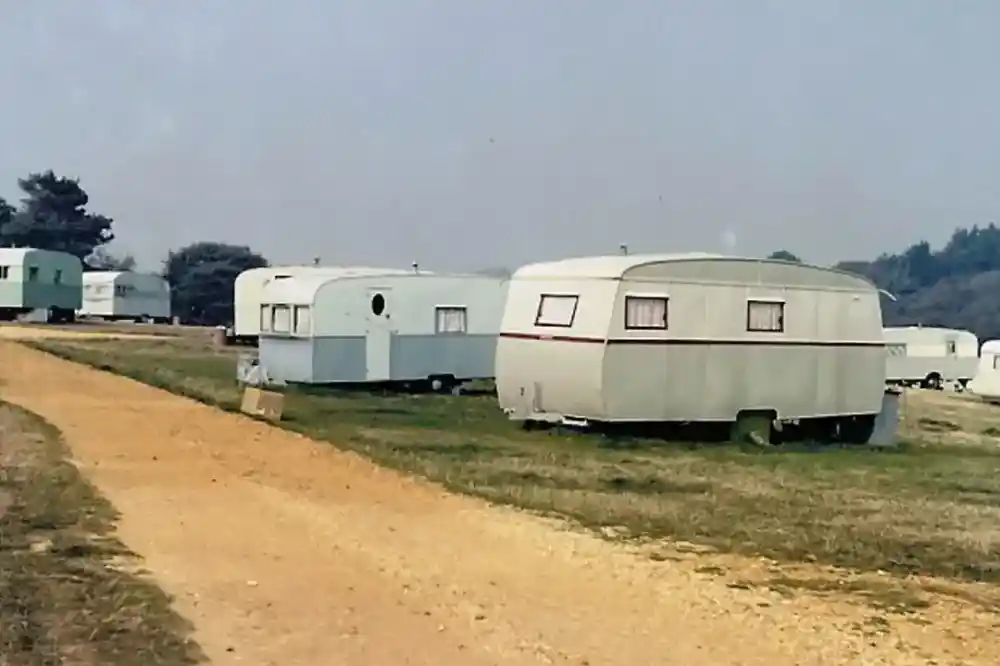A dirt path leads through a grassy area with several vintage travel trailers parked on either side. The trailers have rounded shapes and large windows, reflecting a retro design. Trees can be seen in the background, with a hazy sky overhead.