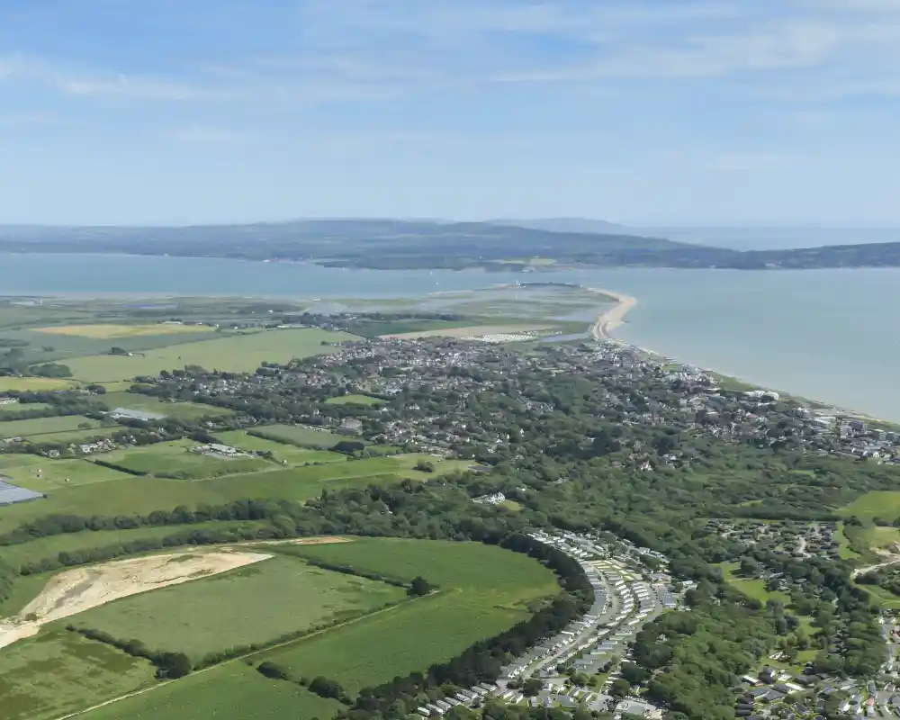 Aerial view of a coastal area with green fields, a sprawling campsite featuring rows of caravans, and distant hills meeting the shoreline. The ocean is visible along the horizon, with a sandy beach curving along the coast.
