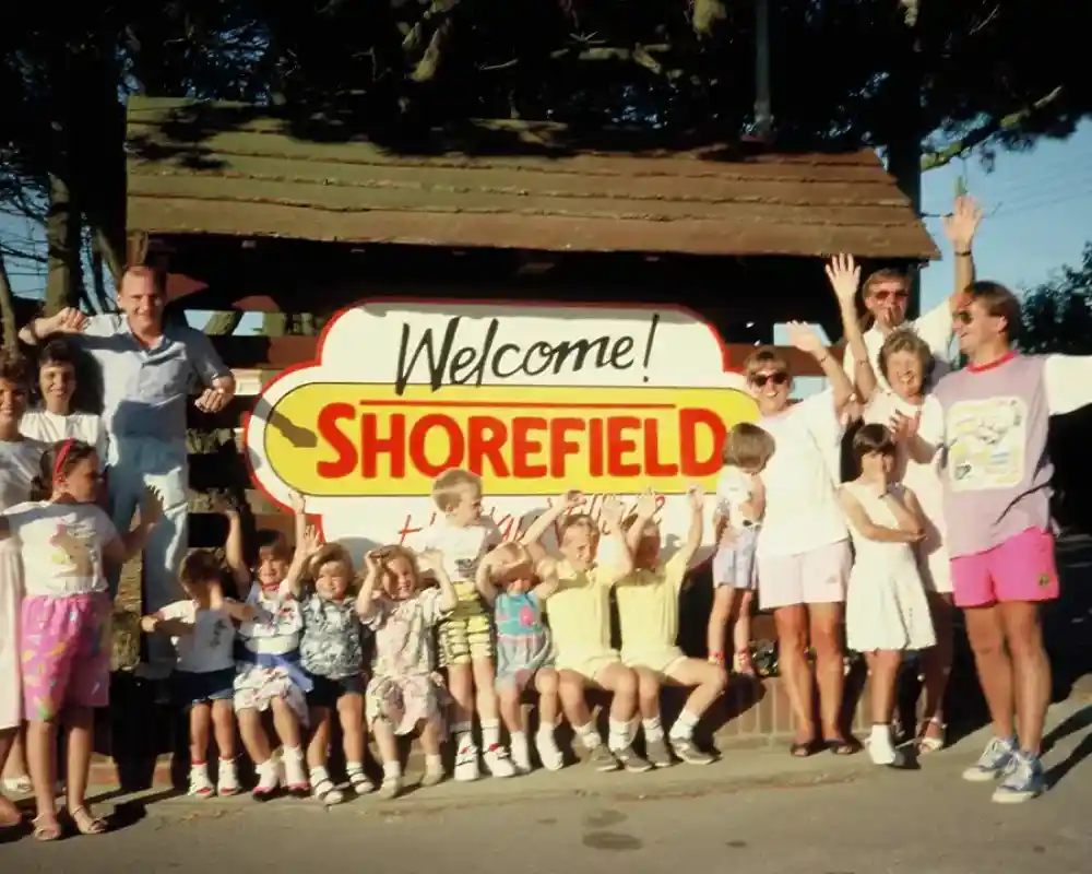 A large group of people, including children and adults, gather around a colorful "Welcome to Shorefield" sign. Many are smiling and waving, exuding a festive atmosphere. The background features trees and a clear sky.