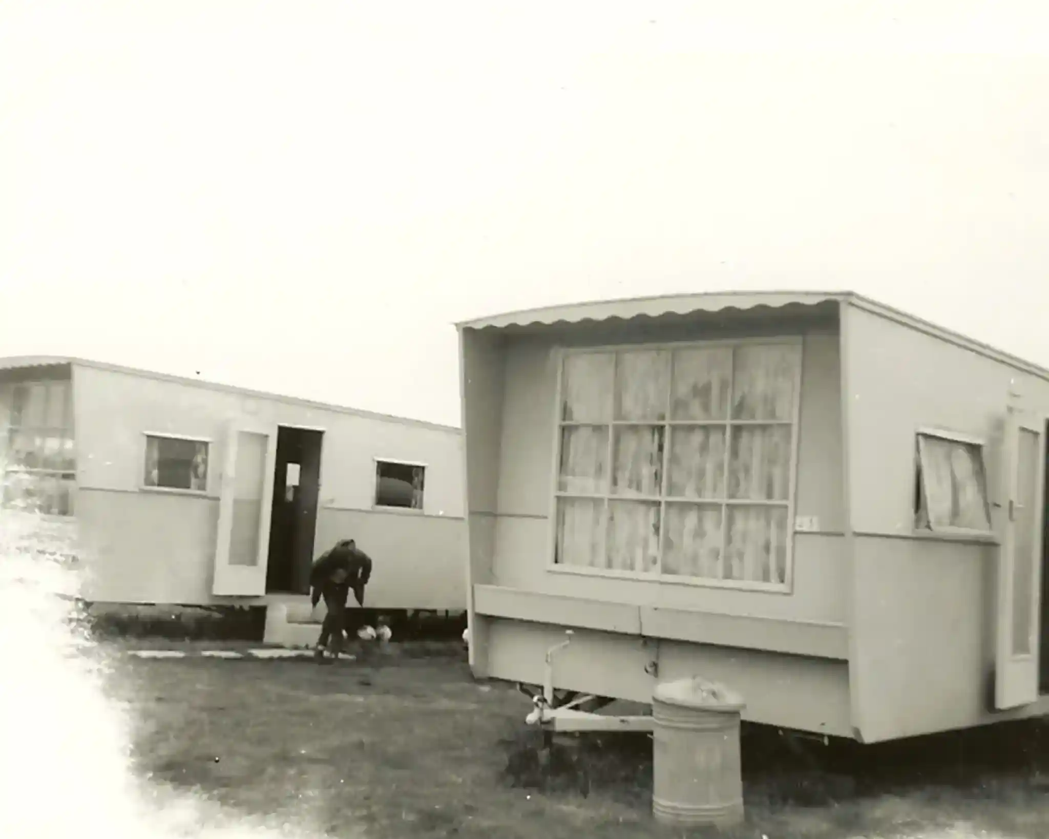 Two vintage mobile homes are parked on a grassy area, with one home featuring large windows and a door open. A person is leaning down near the base of the second home, possibly attending to a pet. The scene is in black and white, evoking a nostalgic atmosphere.
