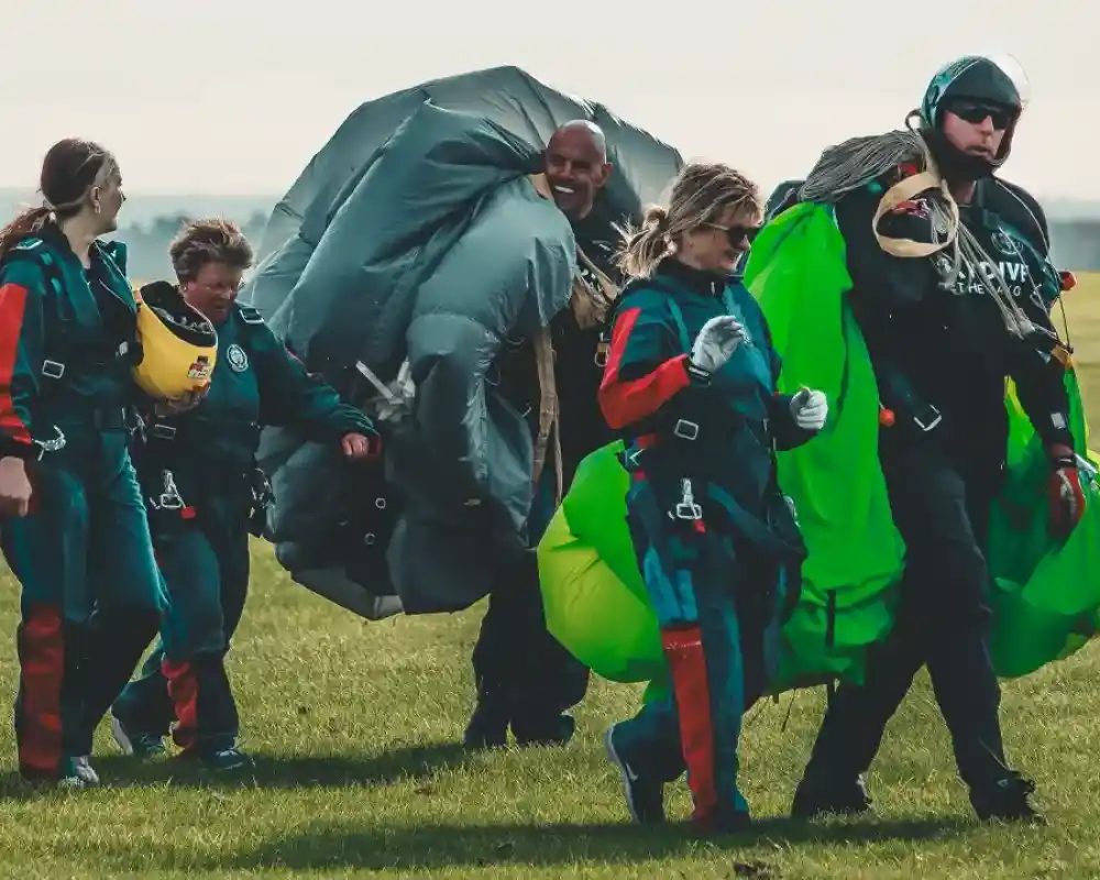 A group of skydivers, dressed in colorful jumpsuits and harnesses, walk across a grassy field. They are carrying parachutes, and some appear to be smiling and chatting with each other. The background features a clear sky and distant hills.