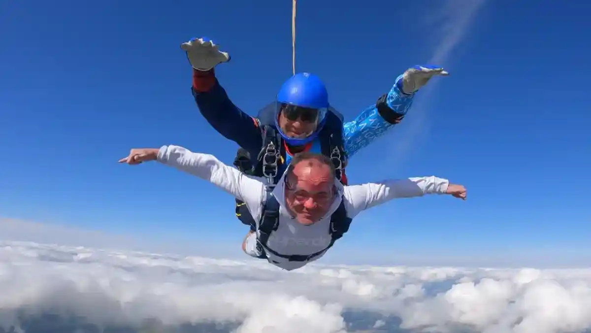 A group of six people stands in front of a car with a green and blue design, holding trash bags and litter pickers. They are smiling and wearing casual clothes, posing outdoors near trees and buildings.