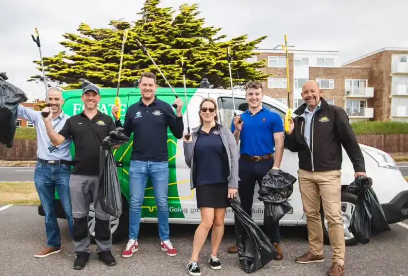 A group of six people stands in front of a car with a green and blue design, holding trash bags and litter pickers. They are smiling and wearing casual clothes, posing outdoors near trees and buildings.