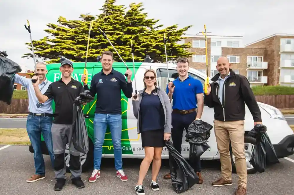 A group of six people stands in front of a car with a green and blue design, holding trash bags and litter pickers. They are smiling and wearing casual clothes, posing outdoors near trees and buildings.