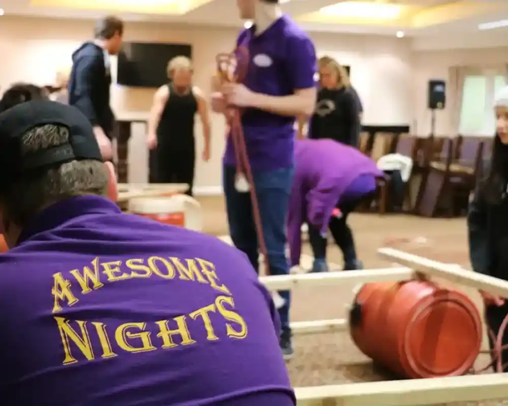 A group of people is engaged in a team-building activity indoors, with some working on assembling a structure. A person in the foreground wears a purple shirt with Awesome Nights printed on the back.
