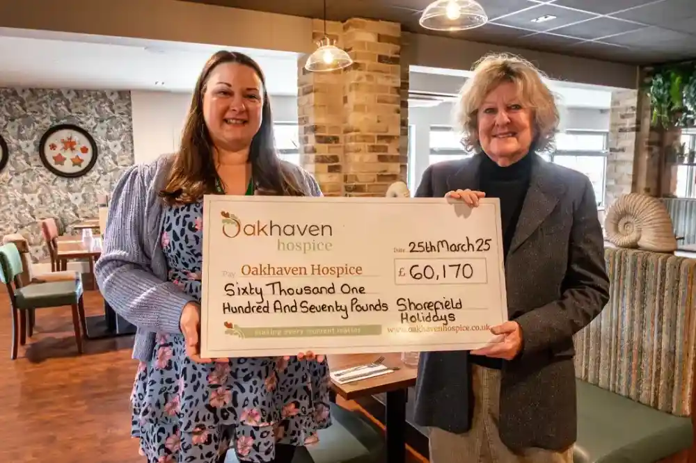 Two women stand in a cafe, holding a large check for £60,170 made out to Oakhaven Hospice. The check is dated March 25. The background features a rustic stone wall and comfortable seating.
