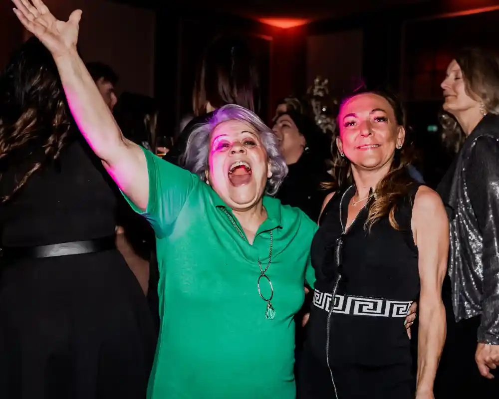 Two women joyfully celebrate at a lively party, one wearing a green dress with her arms raised and a big smile, the other in a black outfit. The background features blurred partygoers, colorful lighting, and a festive atmosphere.
