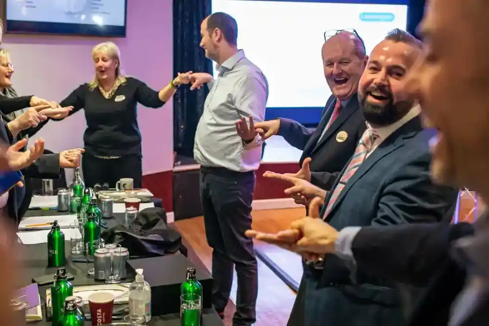 A group of professionals stands in a conference setting, smiling and interacting enthusiastically. Some individuals are gesturing with their hands, while others appear to be engaged in a lighthearted activity. Tables in the foreground are set with green bottles and glasses.