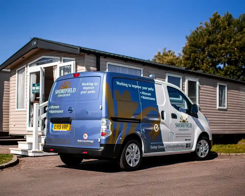 A parked van with Shorefield Concessions branding is next to a mobile home. The van features text about improving parks and mentions eco-friendly practices. Trees are visible in the background, indicating a park-like setting.