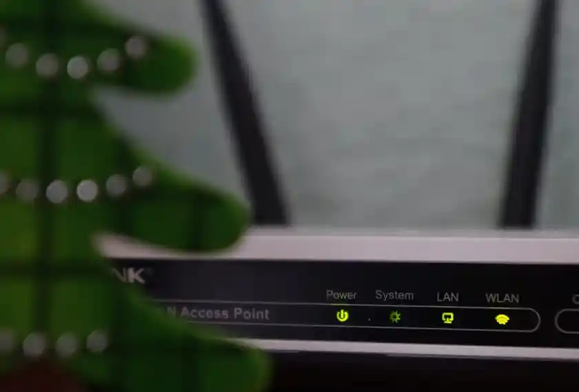 Front view of a WiFi router with indicator lights for power, system, LAN, and WLAN turned on, partially obscured by a decorative green object resembling a Christmas tree