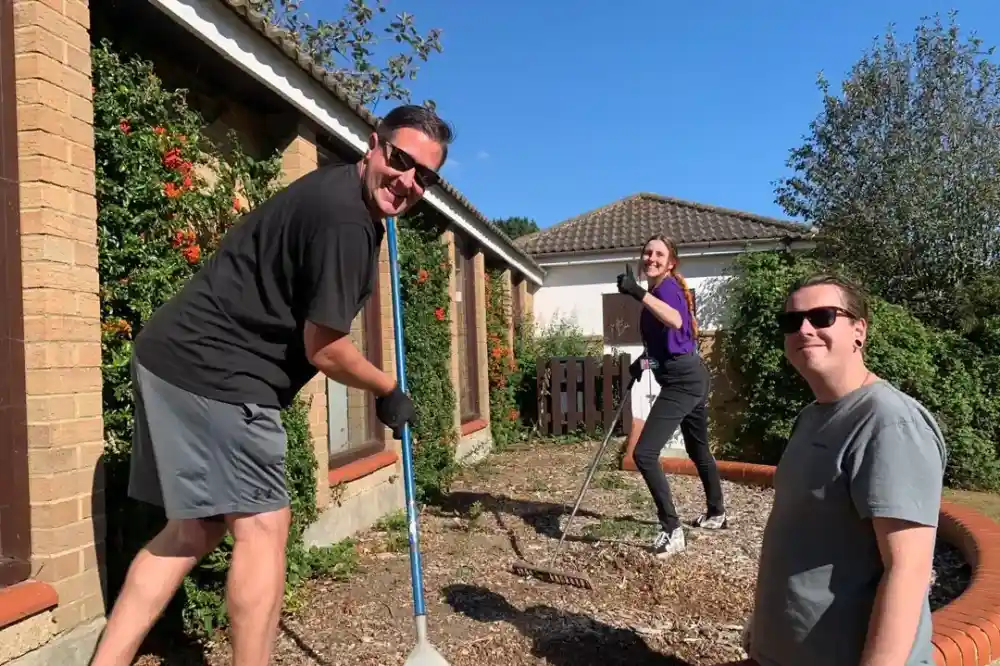 Three people are gardening outside a house. One person is smiling while holding a rake, another is using a hoe, and the third is standing with a trowel. Bright sunlight and greenery surround them.