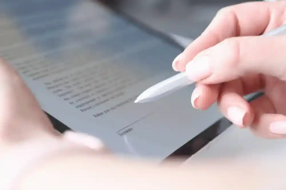 A hand holding a stylus is poised above a touchscreen tablet displaying a document ready for signature.