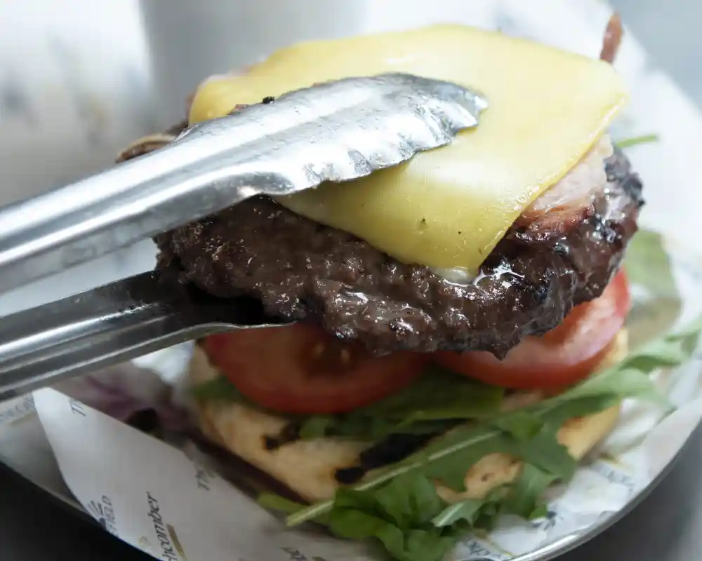 A delicious cheeseburger with a juicy beef patty, melted cheese, sliced tomatoes, and greens on a toasted bun, alongside a cup of crispy French fries. Tongs are holding the cheeseburger, ready to serve.