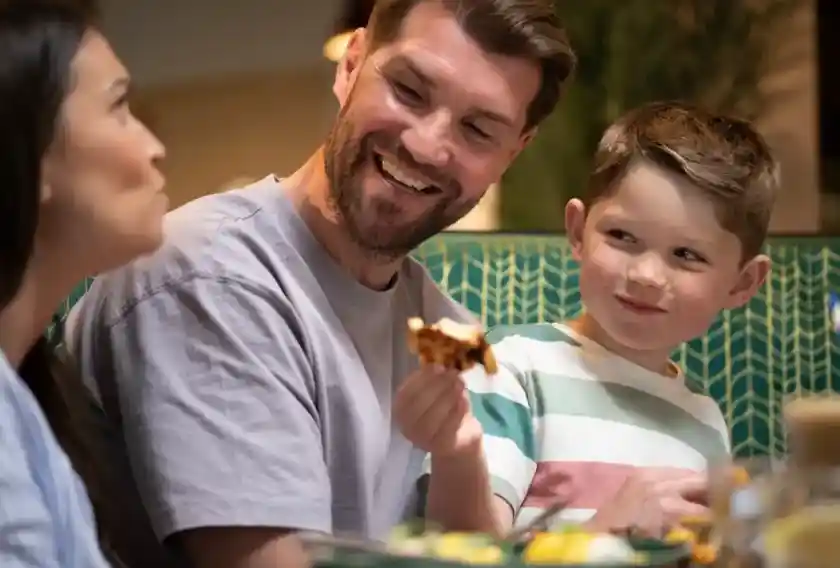 A man smiles while sitting at a table with a woman and a young boy. The boy holds a piece of food, looking playfully at the woman. Brightly colored dishes are spread across the table.