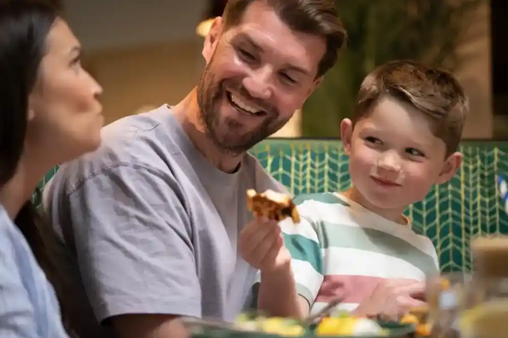 A man smiles while sitting at a table with a woman and a young boy. The boy holds a piece of food, looking playfully at the woman. Brightly colored dishes are spread across the table.