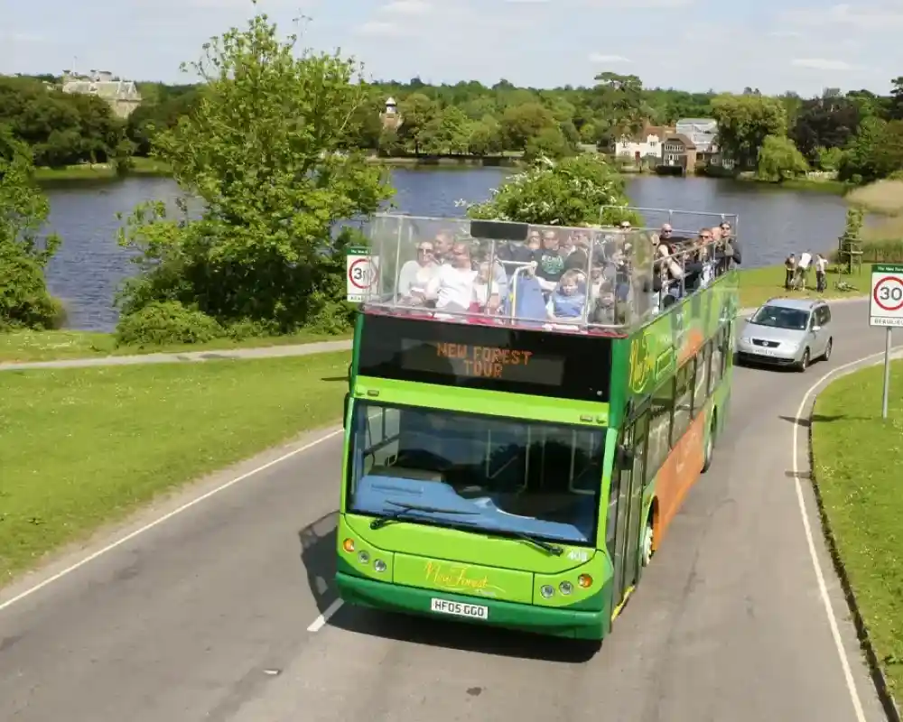 A green and orange double-decker bus with an open-top is driving along a road next to a lake. Passengers are enjoying the view as trees and houses are visible in the background under a clear blue sky.