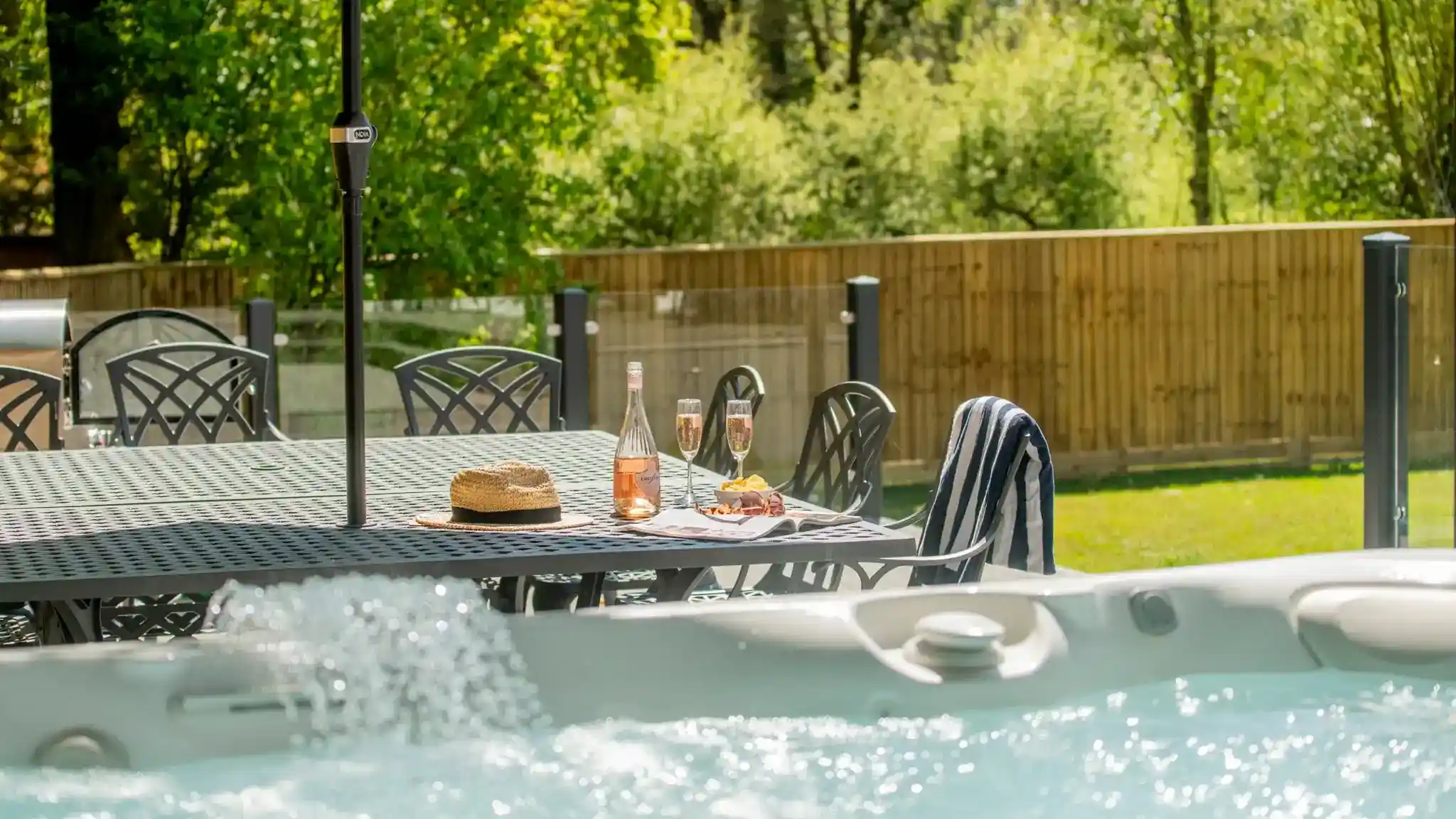 A relaxed outdoor setting featuring a patio table with a bottle of rosé, two champagne flutes, and a plate of snacks. There is a straw hat on the table, and a hot tub is visible in the foreground, with greenery and a wooden fence in the background.
