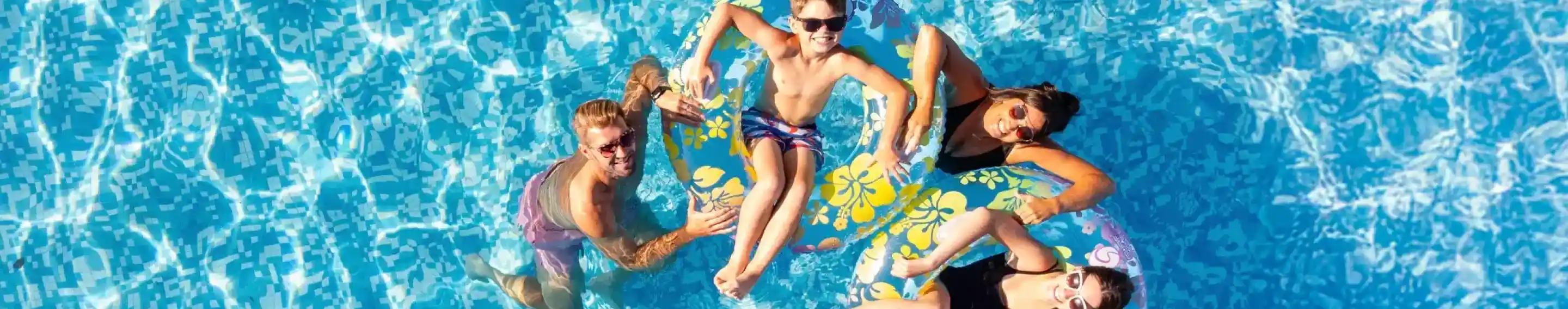 Four people float on colorful inner tubes in a clear blue swimming pool. Two of them are sitting on the tubes, while one is swimming nearby and another leans on the edge. The sunlight creates sparkling reflections on the water's surface.