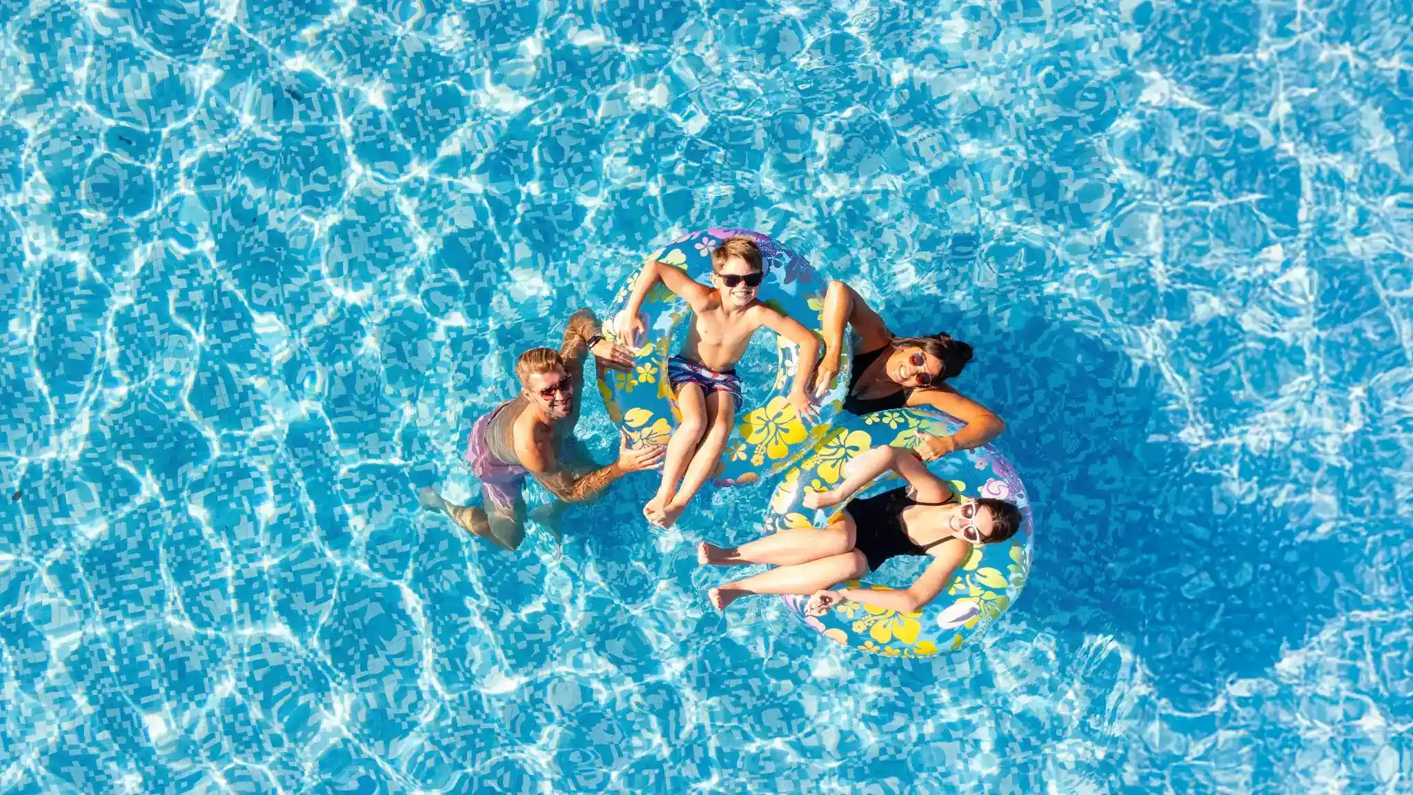 Four people float on colorful inner tubes in a clear blue swimming pool. Two of them are sitting on the tubes, while one is swimming nearby and another leans on the edge. The sunlight creates sparkling reflections on the water's surface.