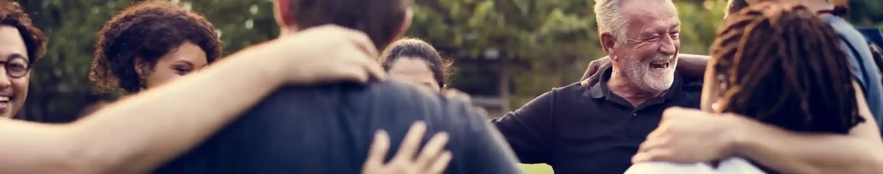 A diverse group of people joyfully embracing each other in a circle outdoors, with smiles and laughter as they connect.