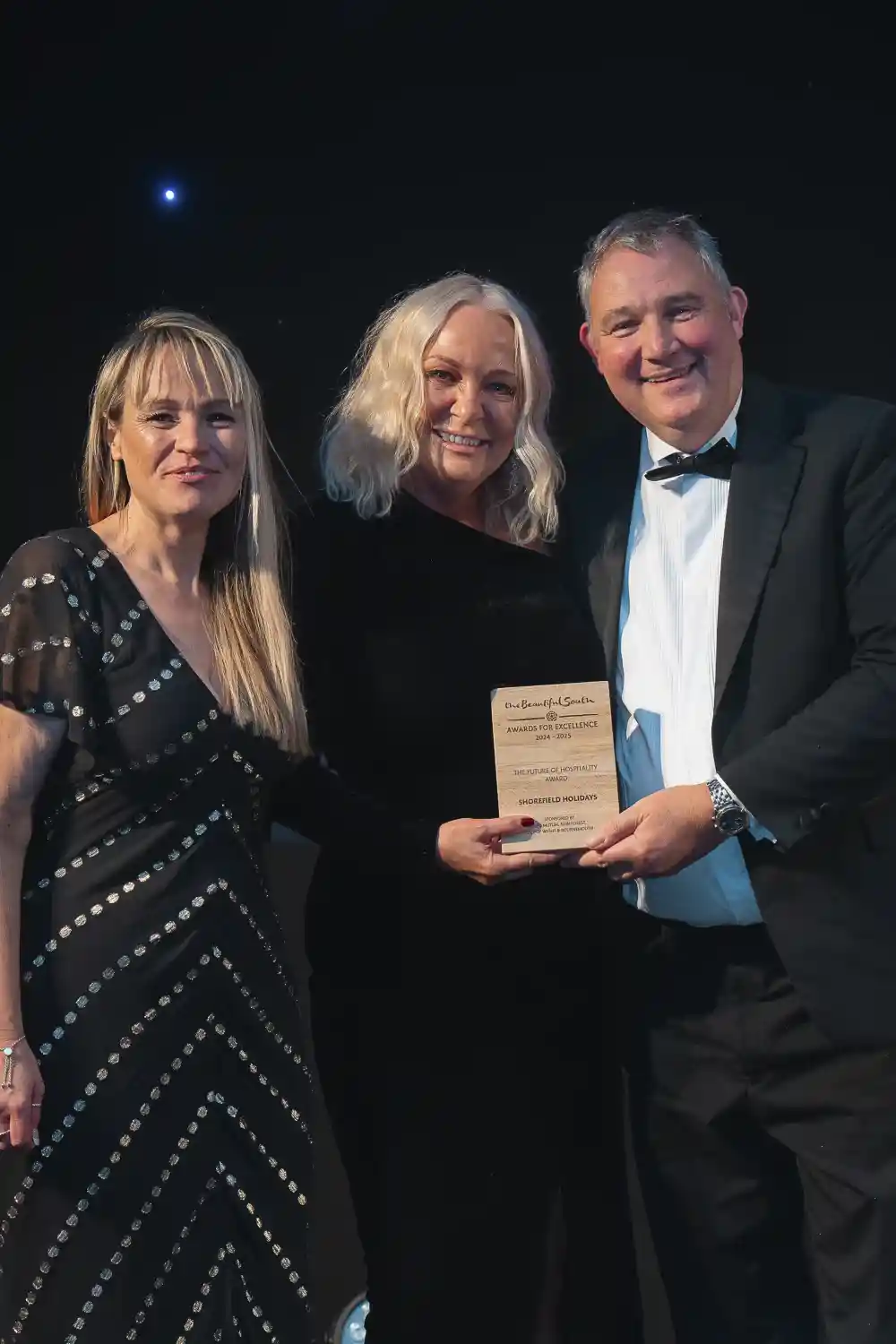 Three individuals pose together at an award ceremony. One woman, wearing a black dress with white polka dots, stands on the left, while a second woman in a black outfit holds a plaque in the center. A man in a tuxedo stands on the right, smiling widely. The backdrop is decorated with subtle lighting.