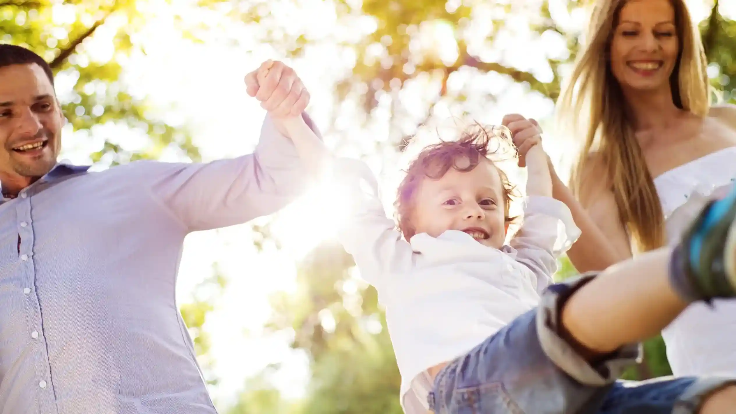 A joyful child swings between two smiling adults, جميعهم holding hands and enjoying a sunny day in a park with green trees in the background.