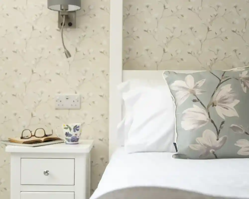 A neatly made bed with white pillows and a patterned throw, next to a white bedside table. The table holds a pair of glasses and a decorative cup, against a floral wallpaper background. A wall-mounted lamp is above the bedside table.