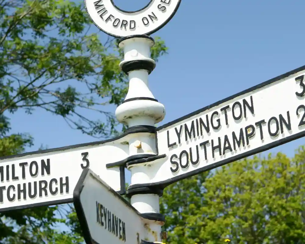 Signpost indicating directions and distances to Milton, Lymington, Southampton, and Keyhaven, surrounded by colorful flowers under a blue sky.