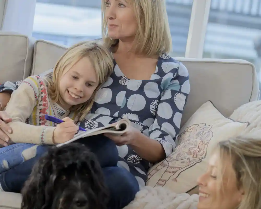 A woman sits on a couch, smiling while a young girl leans against her, drawing in a notebook. Another woman sitting nearby looks on with a smile. A black dog lies on the floor, adding warmth to the cozy, bright living room scene.