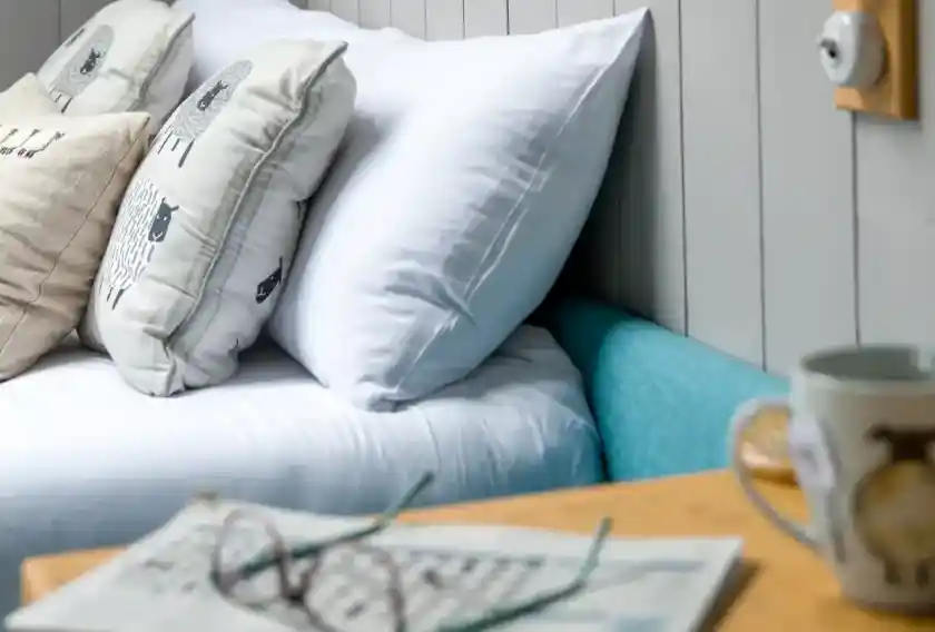 A cosy bedroom corner featuring a bed with white linens and decorative pillows with sheep designs. A wooden bedside table holds a newspaper and a mug. Glasses rest on the table, adding to the relaxed atmosphere.