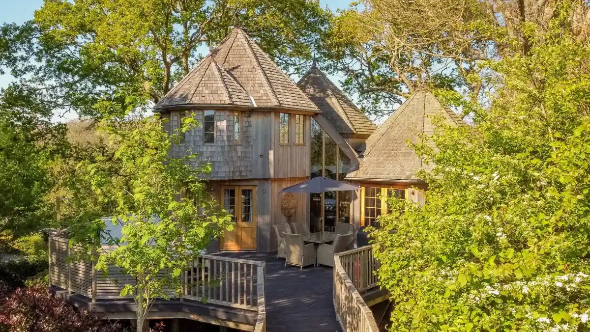 A cosy bedroom corner featuring a bed with white linens and decorative pillows with sheep designs. A wooden bedside table holds a newspaper and a mug. Glasses rest on the table, adding to the relaxed atmosphere.
