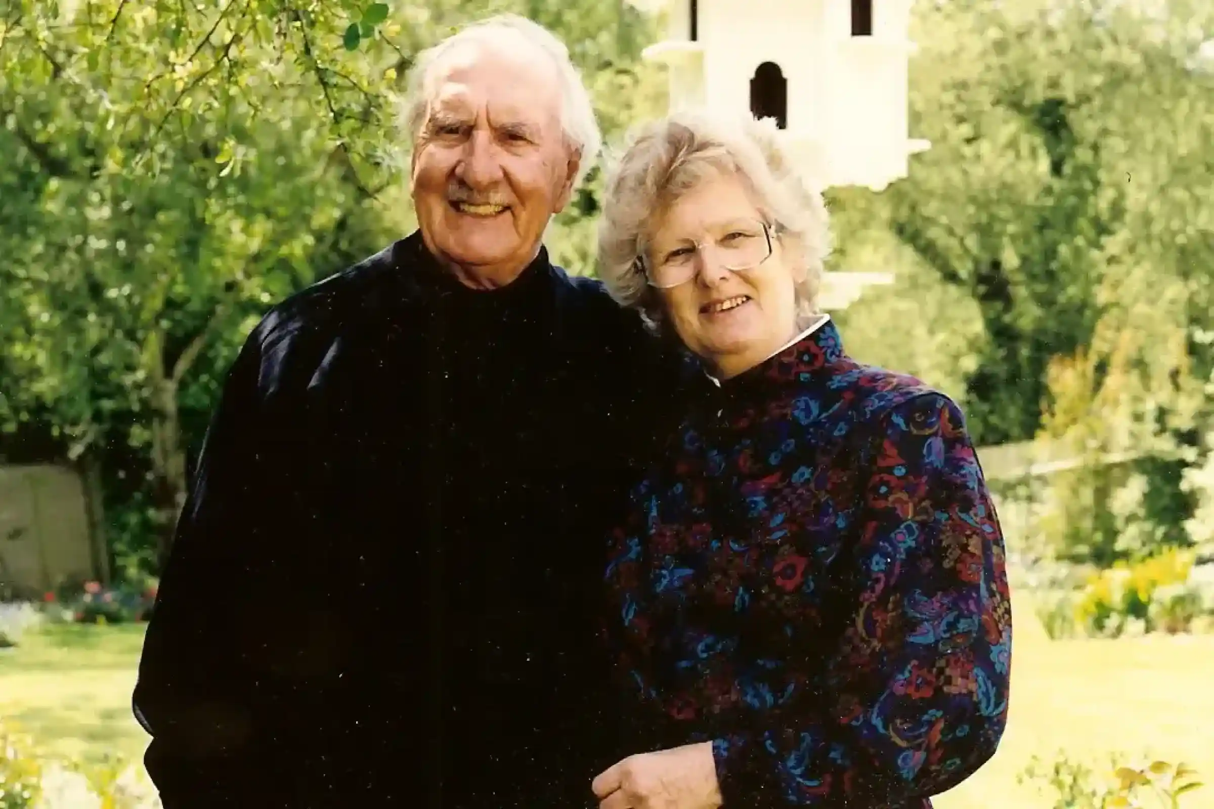 An elderly couple smiles together in a lush garden. The man wears a dark coat and has a cheerful expression, while the woman, in a colourful floral blouse, stands close to him, looking happy. A birdhouse and greenery are visible in the background.