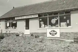 A black and white photograph of a building labeled "Shorefield Caravan Park." The structure features a front door, windows, and signage advertising the park. A flowerbed with gravel is visible in front of the building.