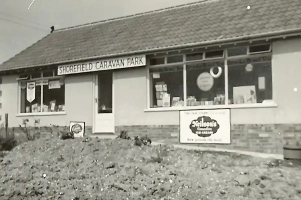 A black and white photograph of a building labeled "Shorefield Caravan Park." The structure features a front door, windows, and signage advertising the park. A flowerbed with gravel is visible in front of the building.