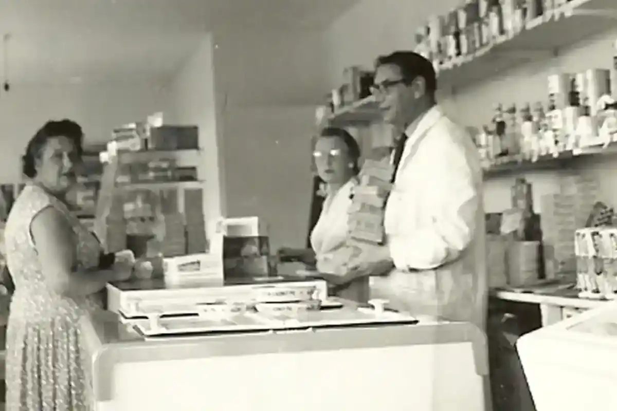 A black and white photo showing a small store interior. A woman in a patterned dress stands at the counter while two shopkeepers, wearing lab coats, assist her. Shelves filled with various products are seen in the background.