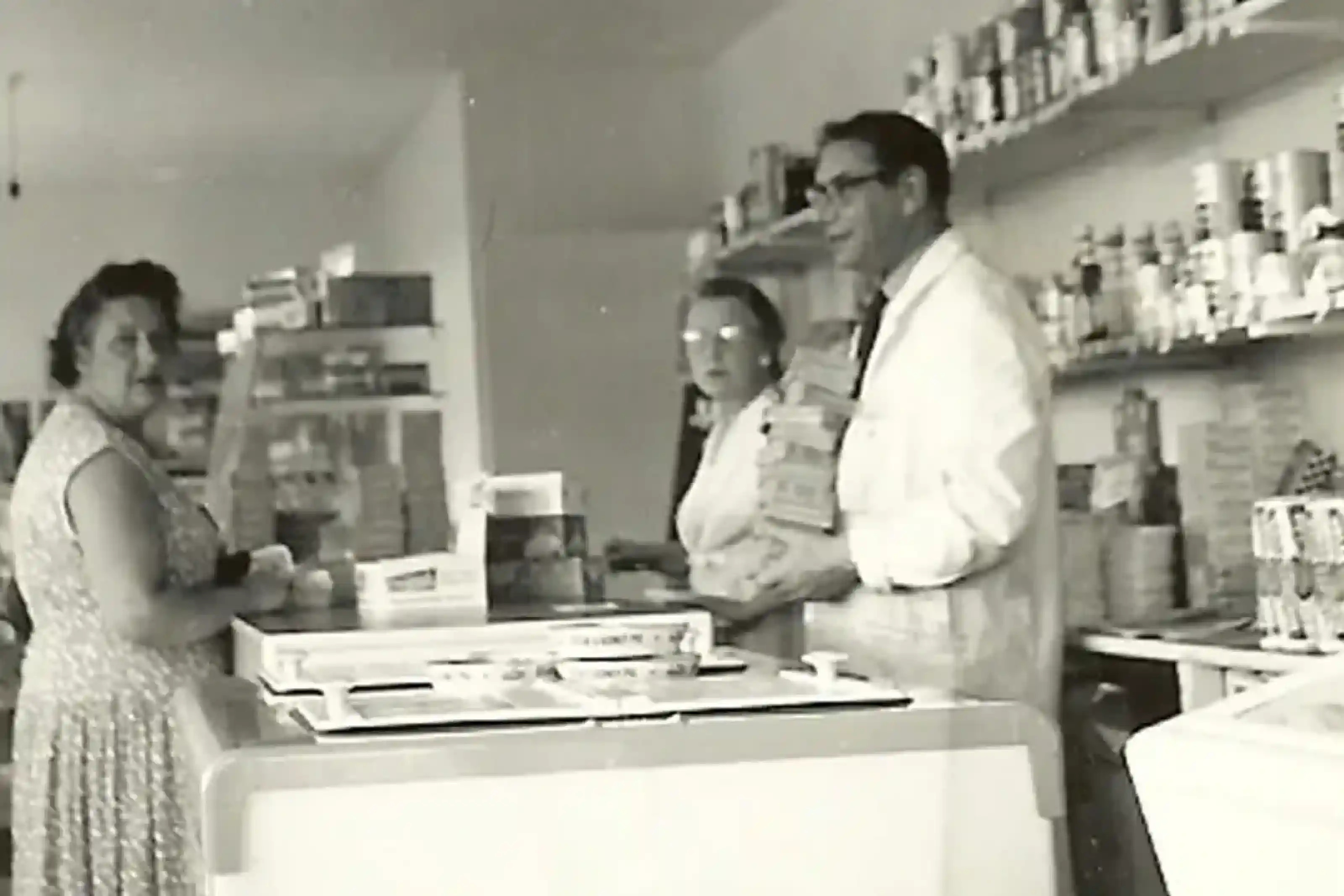 A black and white photo showing a small store interior. A woman in a patterned dress stands at the counter while two shopkeepers, wearing lab coats, assist her. Shelves filled with various products are seen in the background.
