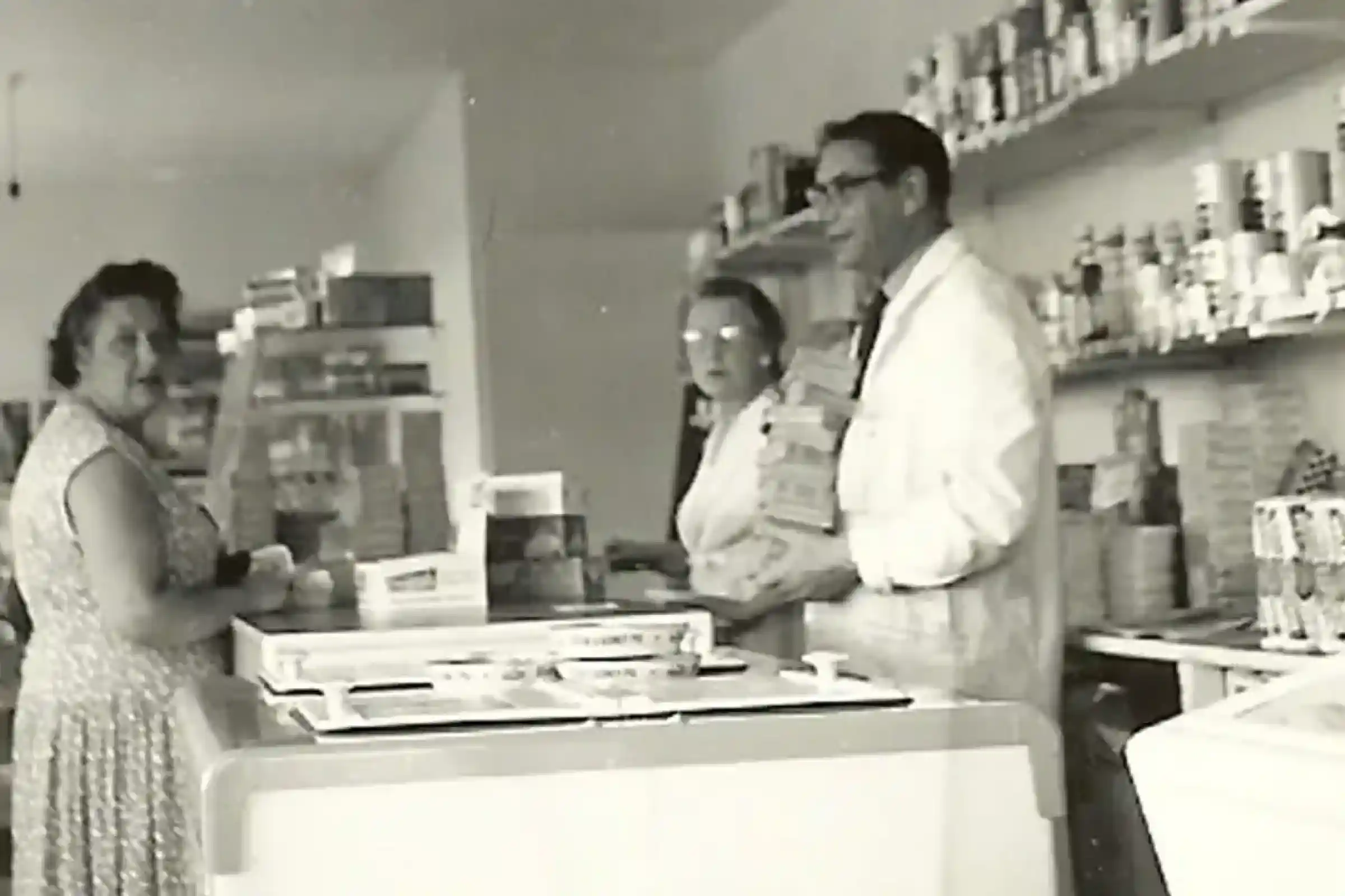 A black and white photo showing a small store interior. A woman in a patterned dress stands at the counter while two shopkeepers, wearing lab coats, assist her. Shelves filled with various products are seen in the background.