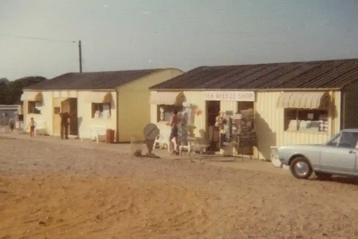 A small vintage storefront with two yellow buildings, featuring a sign for "Sea Breeze Shop." People are outside, browsing or engaging with items displayed. A light-colored car is parked nearby on a gravel surface. The setting appears to be a sunny day.
