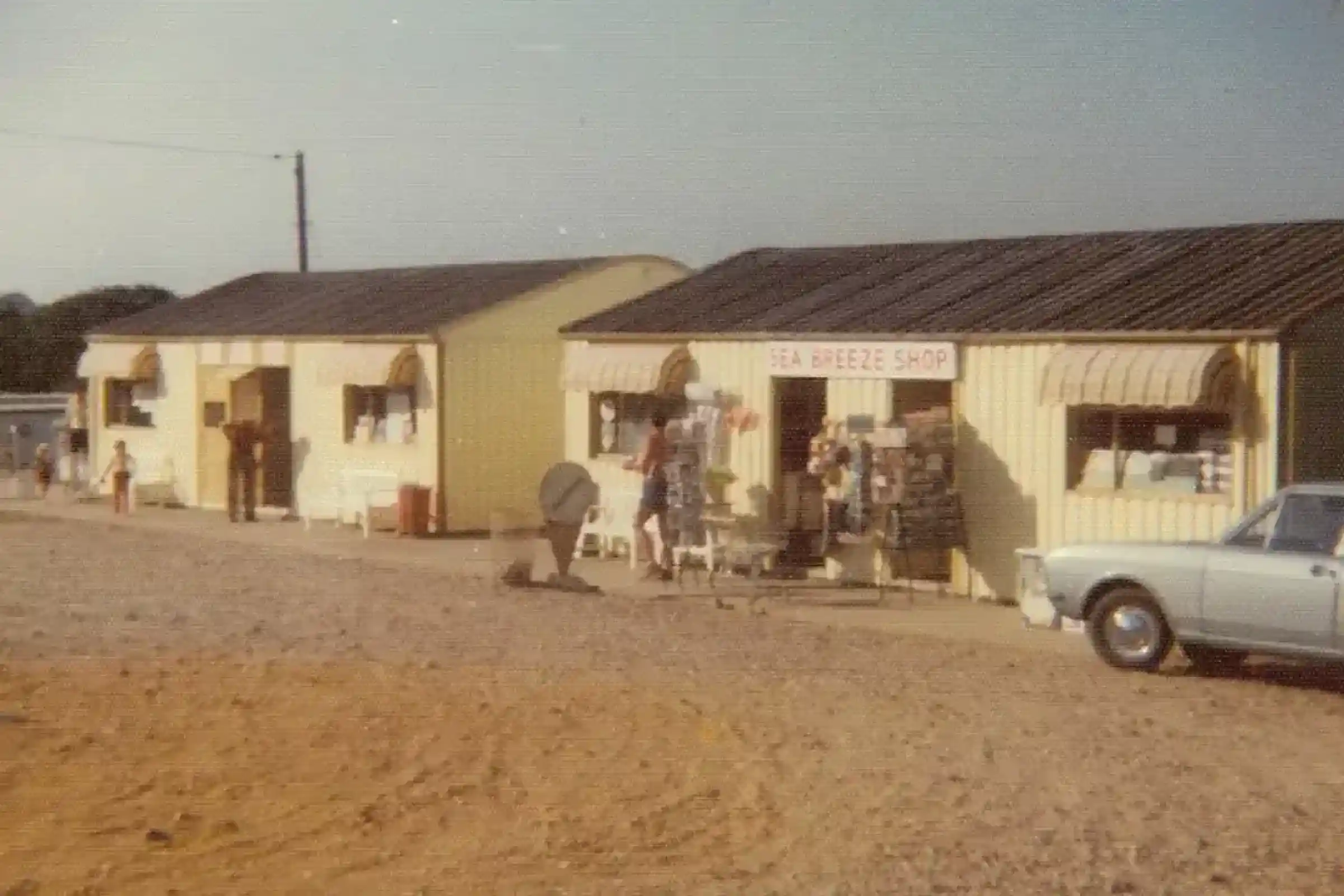 A small vintage storefront with two yellow buildings, featuring a sign for "Sea Breeze Shop." People are outside, browsing or engaging with items displayed. A light-colored car is parked nearby on a gravel surface. The setting appears to be a sunny day.