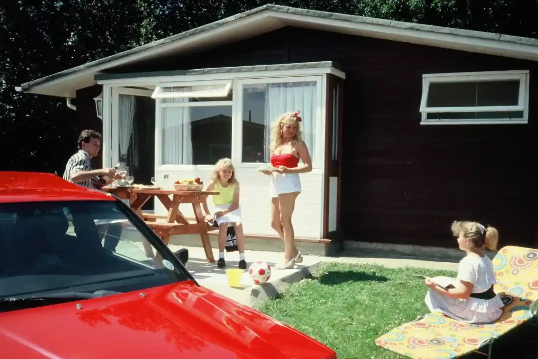 A group of four people enjoying a sunny day outside a cabin. One woman in a swimsuit stands near a picnic table, while another sits on a lounge chair. A red car is parked nearby, and a soccer ball lies on the grass.
