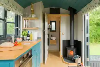 A cozy tiny house interior featuring a kitchen area with a blue cabinet, wooden countertop, and stove. A wood-burning stove is in the corner, and a door leads to a bathroom area with a towel rack. Natural light streams in through the windows, illuminating the vibrant decor.