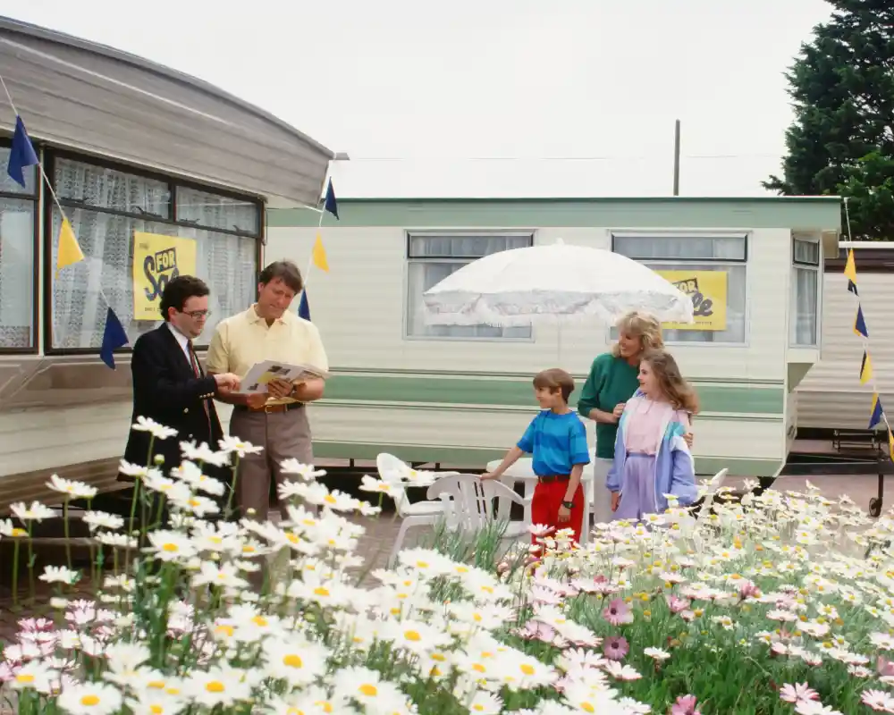 A family stands outside a trailer surrounded by colourful flowers. Two adults and two children engage in conversation, while a woman holds an umbrella nearby. Bunting decorates the trailer, adding a festive touch to the scene.