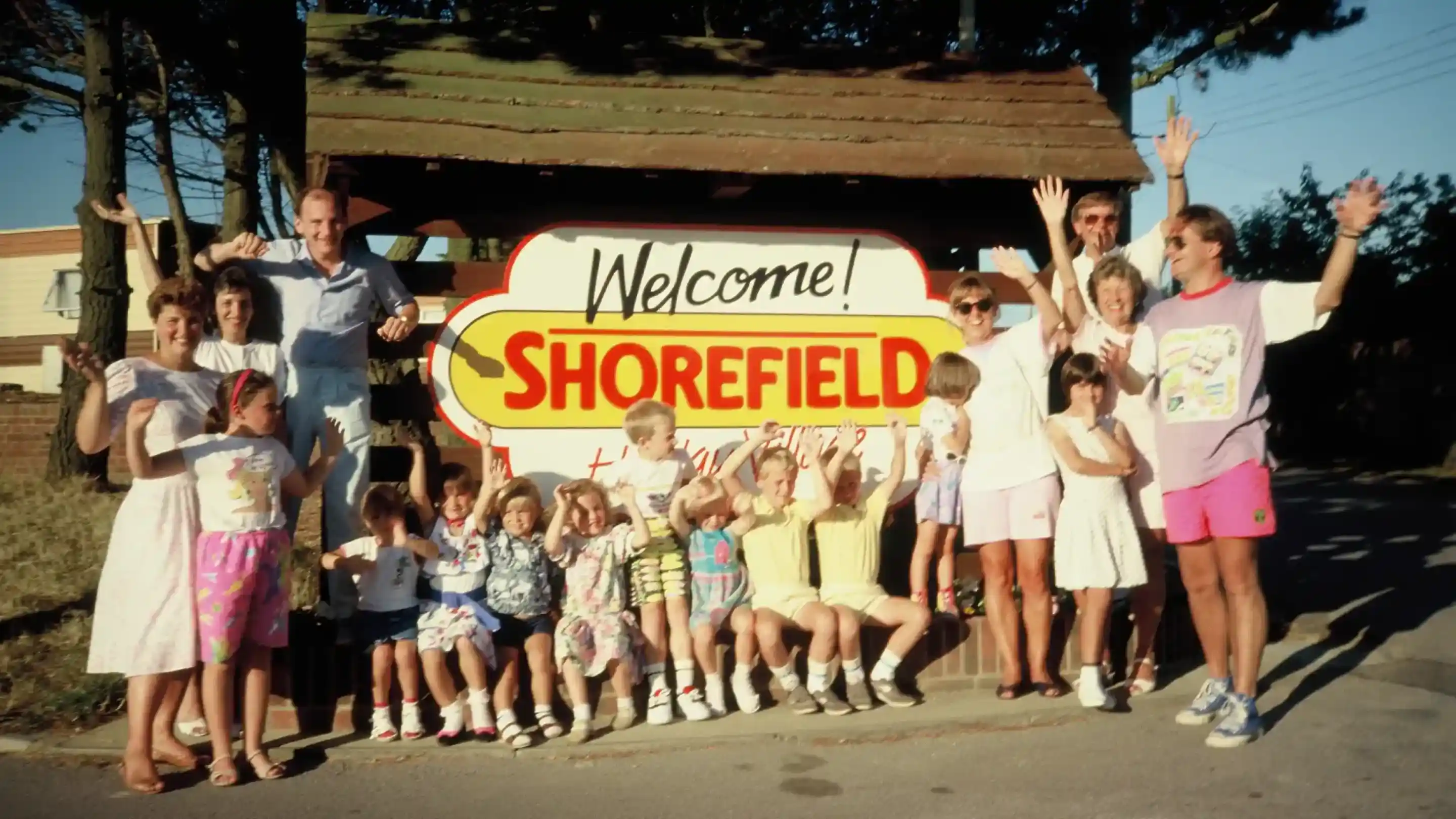 A large group of children and adults joyfully pose in front of a colourful Welcome to Shorefield sign. Everyone is smiling and some are raising their hands, dressed in casual summer clothing. The setting looks like a recreational area or campsite, surrounded by trees.
