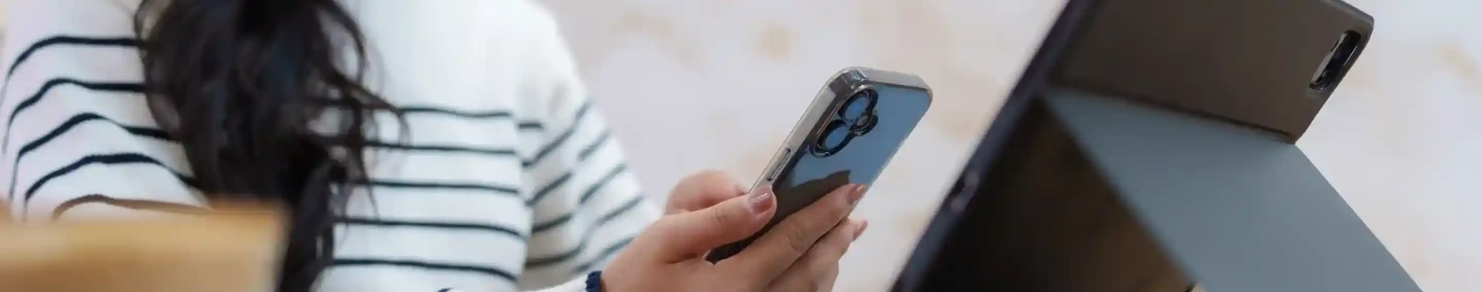 A woman with long, dark hair sits at a wooden table, smiling while holding a smartphone in one hand. In front of her, a tablet is propped up on a stand, and a cup of coffee is visible nearby. She is wearing a striped sweater.