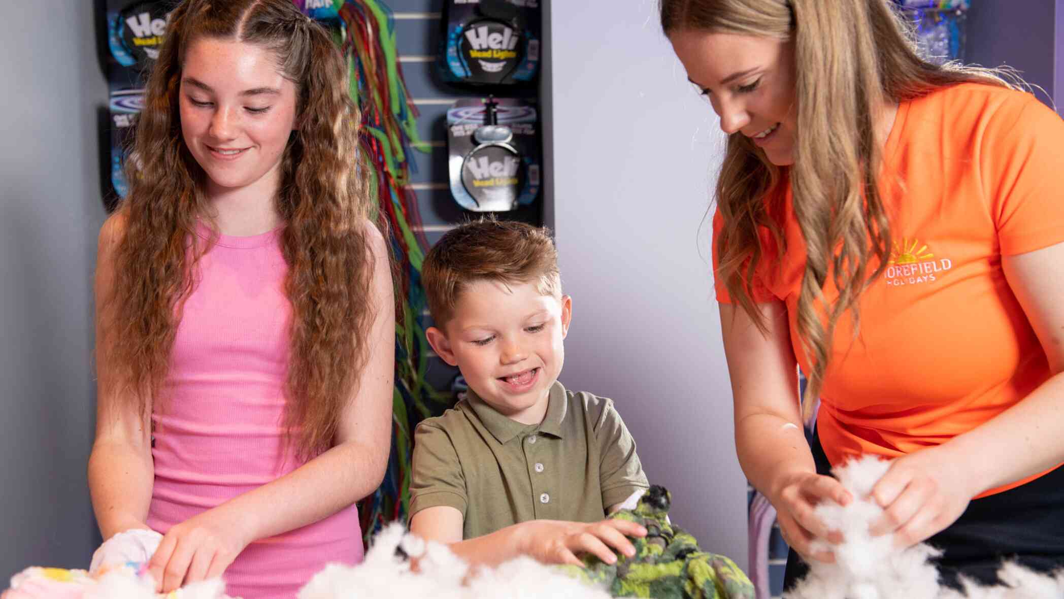 Two children and an adult are crafting together at a table. The girl on the left has long curly hair and is wearing a pink dress. The boy in the center is smiling and wearing a green polo shirt. The woman on the right, dressed in an orange shirt, is assisting them with their project. Colorful materials and supplies are visible on the table.