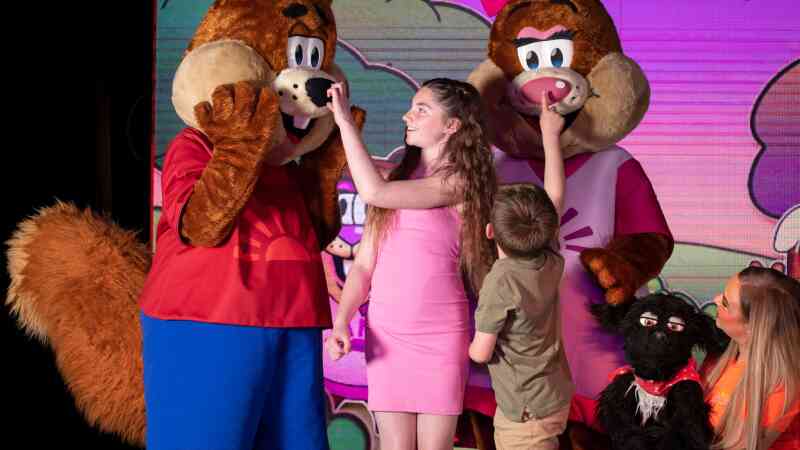 A girl in a pink dress interacts playfully with two mascot characters, one resembling a bear. A boy is leaning in to touch one mascot's nose, while a woman stands nearby, observing with a smile. The background features colorful cartoon graphics.