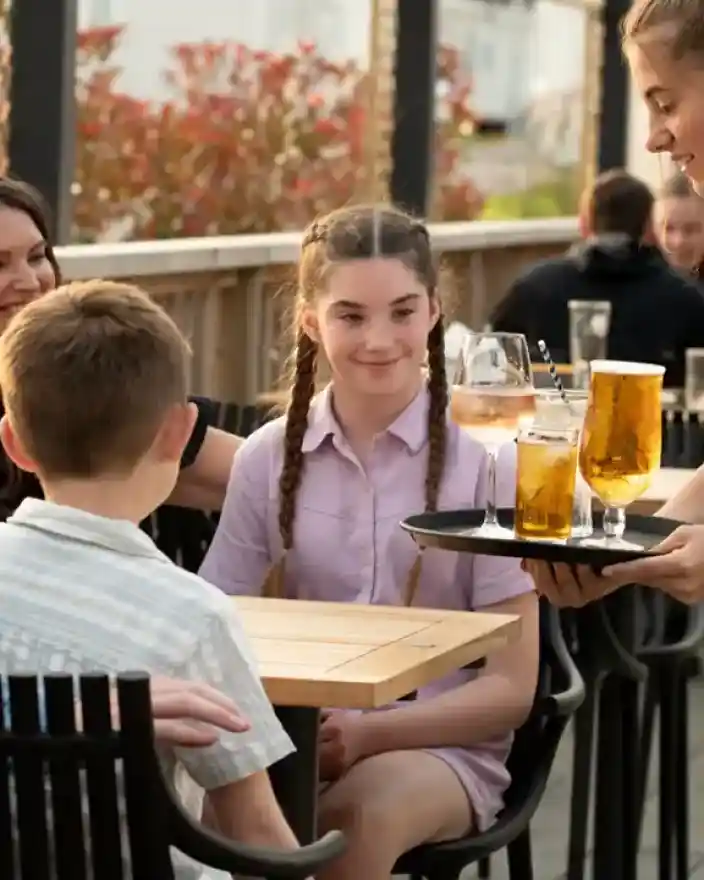 A young waiter serves drinks on a tray to a family seated at an outdoor restaurant table. Two adults and two children are engaged in conversation, with drinks including a large glass of beer and two smaller glasses in front of them. The setting features a patio with greenery in the background.