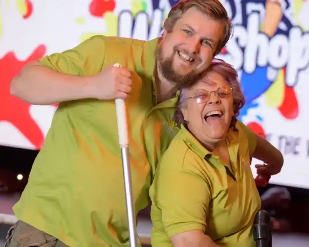 Two smiling individuals wearing matching green shirts pose playfully together. One holds a cleaning tool while the other leans in with a joyful expression. A colorful, cheerful backdrop featuring the words "Workshop" is visible behind them.