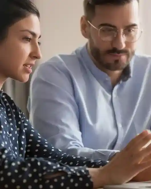 A woman and a man are engaged in a discussion while sitting at a table, with a laptop in front of them. The woman gestures with her hands, while the man listens attentively. A coffee cup is visible on the table.