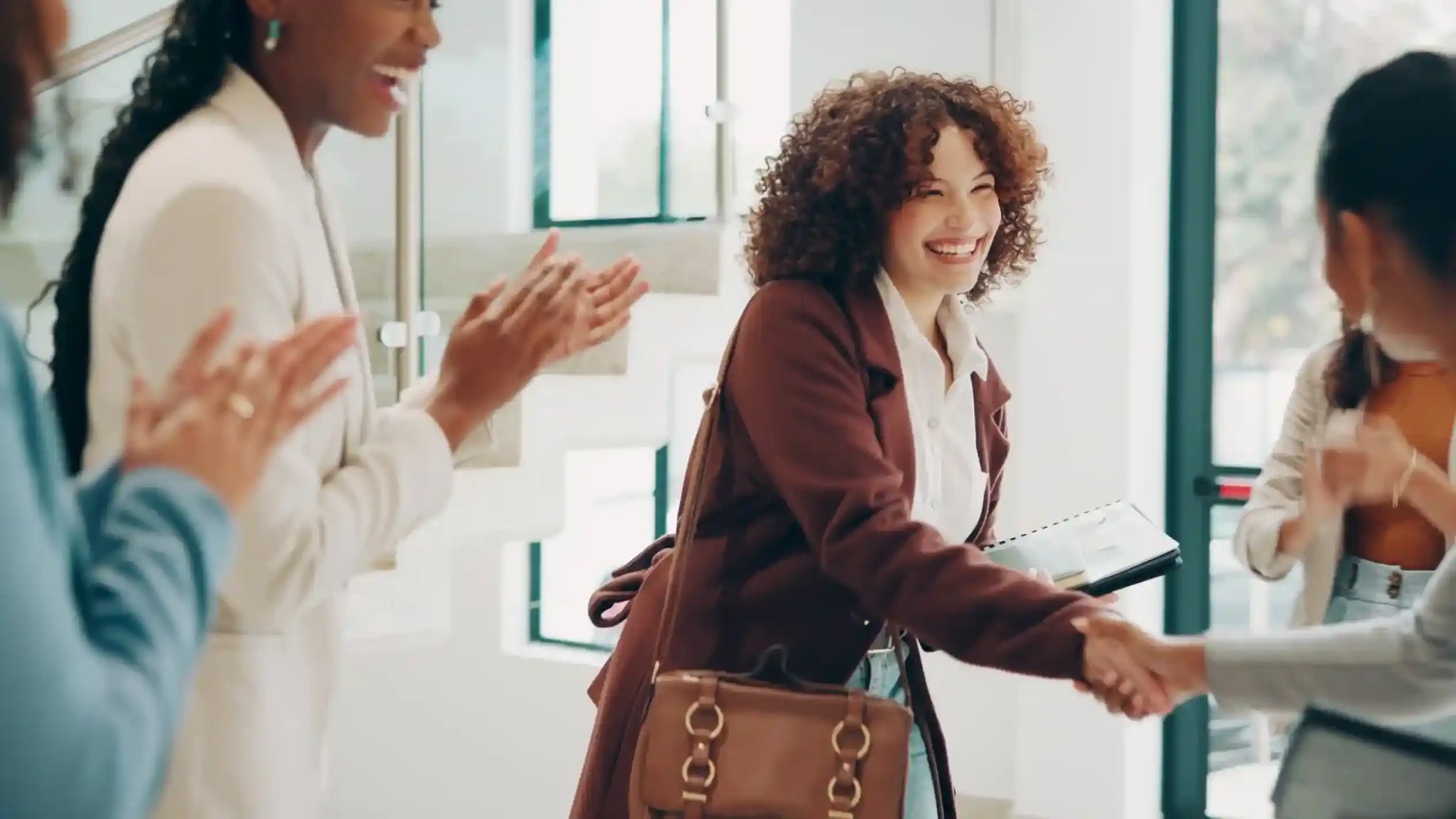 A woman with curly hair smiles while shaking hands with another woman. In the background, a third woman claps, expressing enthusiasm. The setting appears to be a modern, well-lit indoor space.